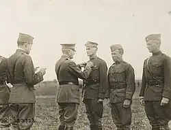 Three men in uniform are standing side by side. The one on the left is wearing a peaked "crush cap" and standing smartly at attention, while the two on the right wear garrison caps and are slouching. A man in a peaked cap and Sam Browne belt is pinning something on the chest of the first man. Behind him stands another man in a garrison cap who is reading a document in his hands.