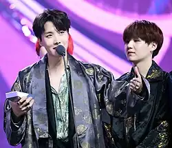 J-Hope, wearing a gray hanbok, speaks into a microphone while holding a Melon Music Award. Suga, on the right side of the image, watches J-Hope speak.
