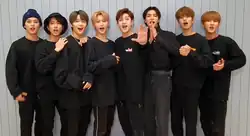 Eight men dressed in black standing closely together in front of a light blue paneled wall.