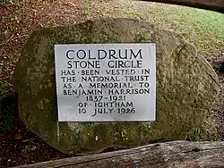 A circular boulder takes up most of the image; behind it can be seen green grass. On the boulder is a light-grey plaque. On this plaque are written the words: "Coldrum Stone Circle has been vested in the National Trust as a Memorial to Benjamin Harrison 1837–1921 of Ightham 10 July 1926". All the letters are in capital letters; those stating "Coldrum Stone Circle" are larger than the others.