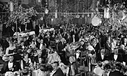 Large, ornate room, filled with people in formal dress sitting at different tables.