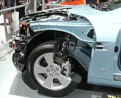 The internal parts of the front left of a light cyan sedan, revealing its engine and some of its wheels and its suspension setup