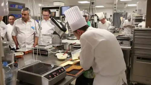 A chef in white clothes pours white chocolate. The room is full of other chefs being observed at their stations.