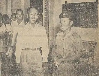 grainy newsprint photo of an Indonesian man standing and smiling in a crowd while a military police officer beside him is also smiling. a group of men standing in the background look unhappy