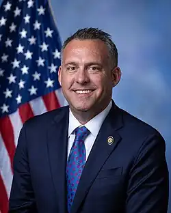 Gray smiling in front of the U.S. flag, wearing a black suit, white shirt, and blue polka dot tie.