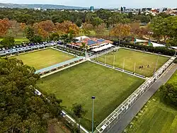 The Adelaide Bowling Club in its present location in the Eastern Parklands, with 3 large playing greens and clubhouse in the foreground and the trees of the parklands and tall city buildings behind.