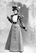 Black-and-white photograph of a young black woman wearing a hat with feathers and a suit: a coat with long, leg-of-mutton sleeves and wide-black lapels with a skirt the same cloth as the coat. The coat's skirt passes just over her hips. The coat is open to show a white blouse beneath. The woman is holding a cane behind her back.