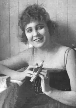 A smiling young white woman with short dark hair, holding a manicure device; she is wearing a slip-style top with bear arms