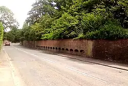 Photograph of a wall with 17 semi-circular grilles built into it. The grilles are spaced horizontally along the wall's length