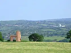 Patchwork of fields and trees with buildings showing in the distance. In the foreground is grass with a ruined building.