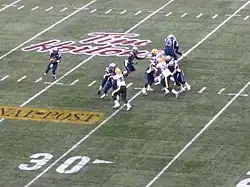 The quarterback, holding the football, looks down field for a teammate to pass to while several of his teammates push up against four opposing players attempting to reach him.