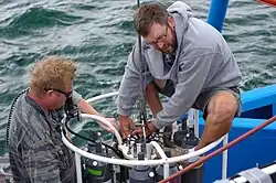 Technicians prepare a CTD-rosette for sampling by pulling back and securing the cables that hold the Niskin bottles in their open positions. When the rosette is in the water, someone on the ship will remotely trigger sampling, and the circular cable holder at the top of the rosette will release cables, closing the bottles one by one at specific water depths.