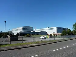 A photograph of a modern factory with two large industrial buildings standing side by side and a loading area at the front. The name of the company is written in red lettering at the front of each building. A column of steam rises in the background