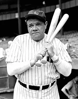 Full body shot of baseball player Babe Ruth, holding a bat and wearing a "NY" hat.