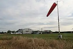 A windsock framing a selection of small buildings on a grass field with several small planes dotted around