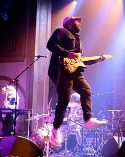 blurry image of Bartees Strange wearing dark clothing and a light-coloured cap, playing guitar mid-leap onstage