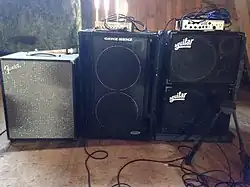 A Fender bass cabinet, a Genz Benz bass cabinet, and two Aguilar cabinets are shown. The Genz Benz and Aguilar cabinets each have amplifier "heads" sitting on top.