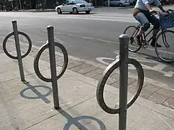 Unpainted steel rings, each welded to a steel post (running through it), set in concrete slabs. A cyclist is passing by from left to right on the cycle path behind.