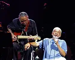 Dupree seated onstage playing guitar with Bill Withers in front of him holding a microphone