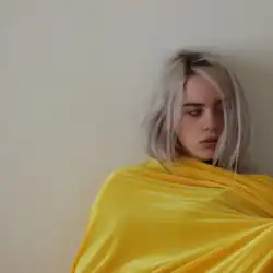 Eilish is seen wearing all yellow, leaning against a wall and looking to the side.