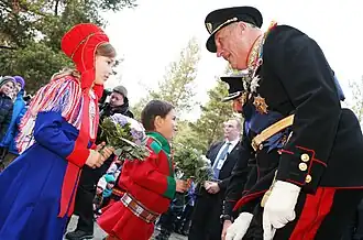 Photograph of the king of Norway speaking to and receiving a bouquet of flowers from a young girl dressed in a traditional Sami gaeptie