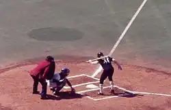A view of home plate from the first base side seats. A left-handed batter with the number 48 on his black uniform is in the batter's box looking down at home plate with one hand on the bat which is resting on his shoulder. The catcher in a white uniform has just caught a ball close to the ground. The umpire, wearing blue pants and a red jacket, is leaning over the catcher.