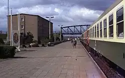 View of a train stopped at a long railway platform, at the end of which is an arched iron bridge. A grey concrete barracks and East German state emblem are visible on the side of the platform. Several people are standing or walking on the platform and the train's doors stand open.