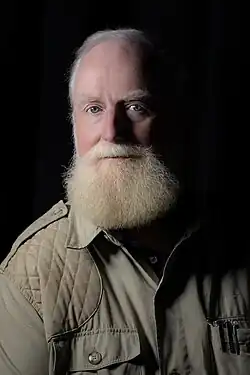 Action portrait of a man in his fifties with a bushy, strawberry-blond beard seated while speaking wearing a safari jacket and gold watch