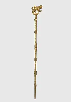 A gold staff topped with a sculpture of a cat and a mouse attempting to crawl inside the cat's bag.