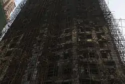 An upward view of a burned-out high-rise: a blackened mess of tangled scaffolding and holes for windows.