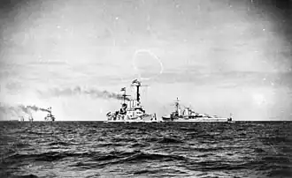 Several light gray warships are seen steaming in the distance; most are steaming generally toward the viewer, while the Königsberg-class cruiser is sailing in the opposite direction.