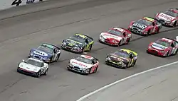 a single road car leads a group of race cars which are being driven in a double-file queue