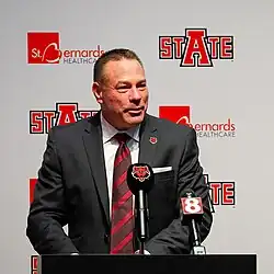 Butch Jones wearing a gray suit and standing behind multiple microphones speaking at a press conference.