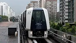 A two-car Mitsubishi Crystal Mover train, numbered 47, is shown on elevated tracks approaching an LRT station in Singapore. The train is white and dark blue, and behind it, a dense urban landscape of tall residential buildings is visible on both sides of the tracks.