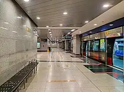 A train arrives at the CCL platform (on the right). On the left is a grey wall with a black rectangular seat engraved with the station's name.