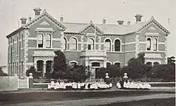 A monochrome image of the Carlile House photographed in 1909, then known as the Richmond Road Children's Home. A large crowd mostly consisting of children are seen dressed in white uniform standing outside the building.