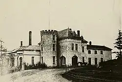 A black and white photograph of a manor, featuring stone towers similar to a European castle. Some pine trees are visible nearby.