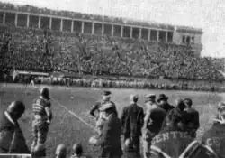 A group of football players standing on the sideline of a football field before a large crowd seated in the stadium