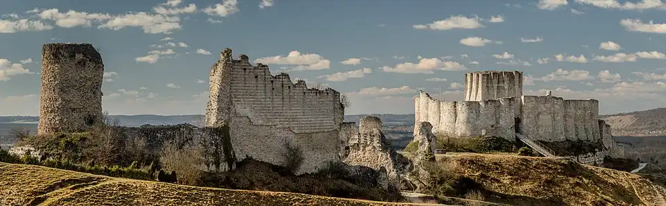 The ruins of a castle in grey limestone dominating the landscape. The River Seine is in the background. The castle's keep protrudes above the walls of the inner bailey on the right, with a bridge leading up to the bailey's entrance. To are ruins of the wall enclosing the outer bailey; a tower stands taller than the ruined walls.