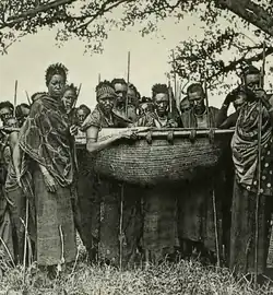 A group of armed warriors stand around a litter, carrying Kabare.