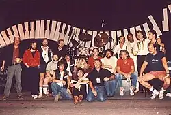 Christine McVie and her touring band posing together on stage, with a giant wave-shaped piano-key prop in the background. McVie is in the middle, surrounded by band members