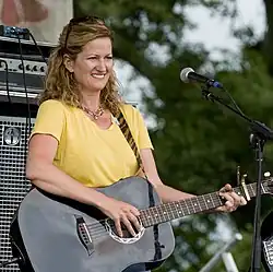 A woman wearing a yellow t-shirt smiling and playing a black acoustic guitar.
