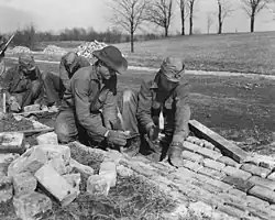 Five workmen. One is holding a shovel, while the other four are laying bricks to form a drainage ditch along the side of a road.