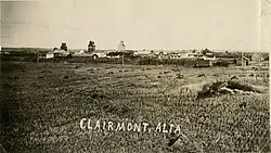 A black and white image of a rural town with three elevators and town buildings from an adjacent hay field