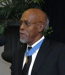 Clarence Sasser, a gray-haired black man standing at a microphone, wearing a medal on a blue ribbon around his neck