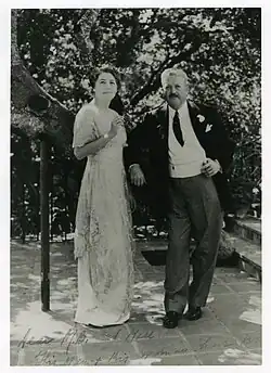 Dr. Logan Clendening stands to the left of his wife, Dorothy Hixon Clendening. He leans on the edge of an invisible object. He is wearing a suit. Dorothy is wearing a white dress and holds a cigarette.