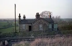A photograph of the now demolished railway station at Constable Burton in Wensleydale, Yorkshire, England. A single railway line runs in front of the building. Two boarded-up windows are shown, as well as an open door which led into the old booking office. Four chimney stacks, some with large ornate Victorian chimneypots are on the roof. A telegraph pole stands to the left of the station.