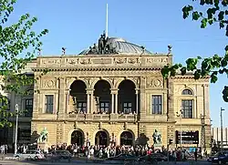 The front facade of a theatre, people entering while others look down from the open balcony