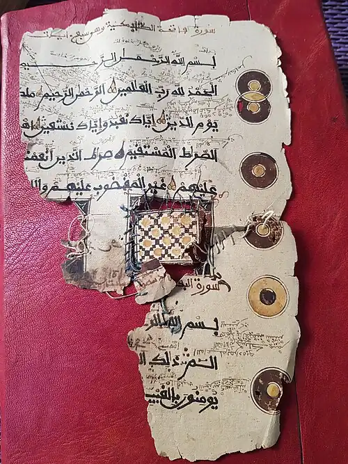 Photograph of an Ancient fragment of a Quran manuscript annotated in Old Kanembu, featuring circular decorative motifs. The document features several circular decorative motifs in brown and yellow along the margins, as well as detailed handwritten commentary in Old Kanembu, inserted interlinearly and in the margins. The parchment shows visible signs of wear and old repairs, with thread stitching running through the paper.