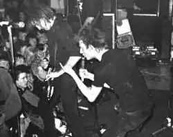 Two members of the rock band Crass are shown at a performance. From left to right are an electric guitarist and a singer. Both are dressed in all-black clothing. The singer is making a hand gesture.